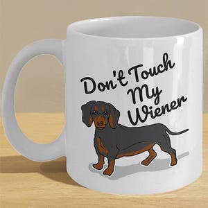 May include: White ceramic coffee mug with a black and brown dachshund illustration. The mug has the text "Don't Touch My Wiener" printed on it.