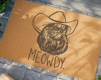 May include: A brown doormat with a black and white illustration of a cat wearing a cowboy hat. The text "MEOWDY" is written below the cat.