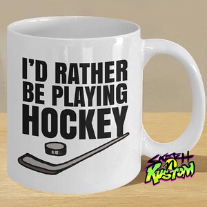 K&ouml;nnte beinhalten: Wei&szlig;e Keramik-Kaffeetasse mit schwarzem Text, der "Ich w&uuml;rde lieber Hockey spielen" lautet, und einer Schwarzwei&szlig;illustration eines Hockeyschl&auml;gers und eines Pucks. Die Tasse hat einen schwarzen Henkel und ein wei&szlig;es Innenleben.