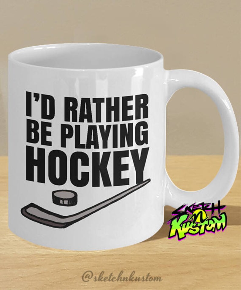 K&ouml;nnte beinhalten: Wei&szlig;e Keramik-Kaffeetasse mit schwarzem Text, der "Ich w&uuml;rde lieber Hockey spielen" lautet, und einer Schwarzwei&szlig;illustration eines Hockeyschl&auml;gers und eines Pucks. Die Tasse hat einen schwarzen Henkel und ein wei&szlig;es Innenleben.