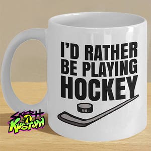 K&ouml;nnte beinhalten: Wei&szlig;e Keramik-Kaffeetasse mit schwarzem Henkel. Die Tasse hat ein schwarzes Motiv mit dem Text "Ich w&uuml;rde lieber Hockey spielen" und einem Hockeyschl&auml;ger und Puck.