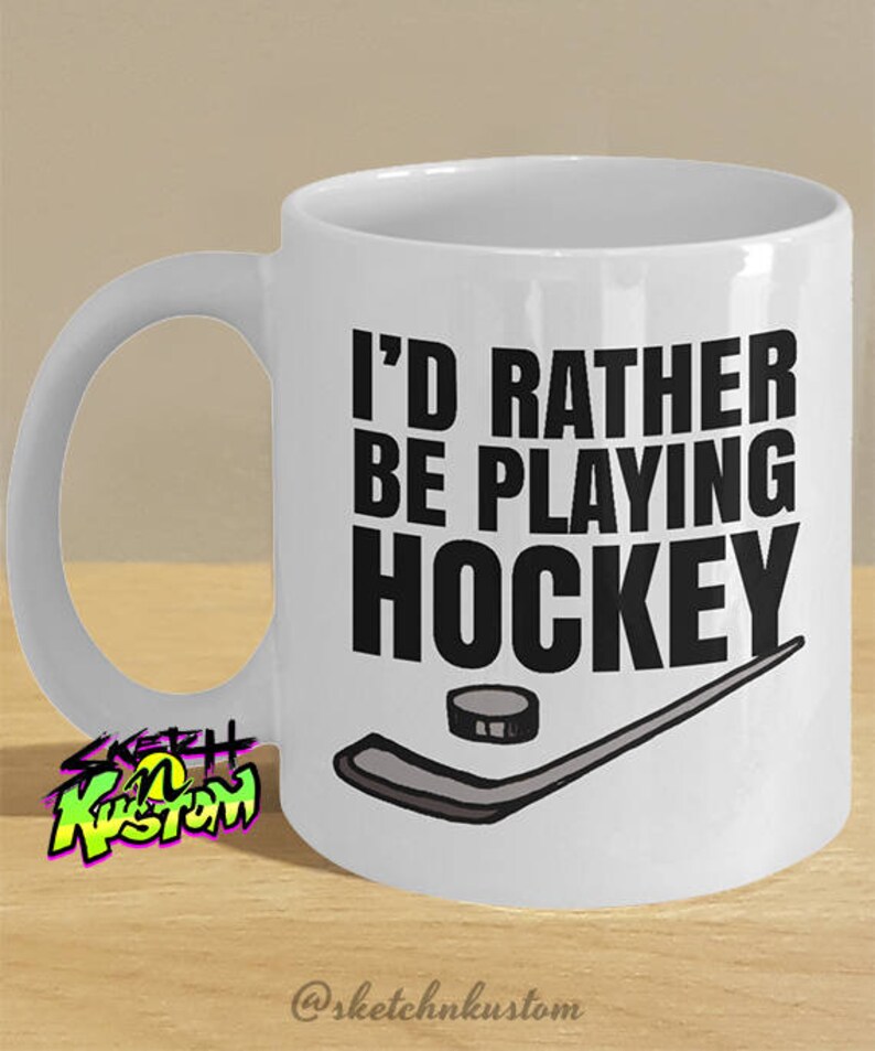 K&ouml;nnte beinhalten: Wei&szlig;e Keramik-Kaffeetasse mit schwarzem Henkel. Die Tasse hat ein schwarzes Motiv mit dem Text "Ich w&uuml;rde lieber Hockey spielen" und einem Hockeyschl&auml;ger und Puck.