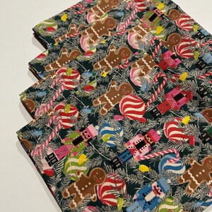 May include: A stack of four folded fabric napkins featuring a dark green background with a repeating Christmas pattern of brown gingerbread people, colorful nutcrackers, red and white candy canes, and multi-colored holiday light bulbs.