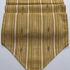 May include: A mustard yellow and white table runner featuring a vertical striped pattern with dashed lines and small grey oval accents. The fabric has a textured weave and the runner is folded into several layers, ending in a pointed triangular edge.