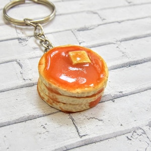 May include: A keychain featuring a miniature stack of pancakes with syrup and a pat of butter. The pancakes are light brown, topped with orange syrup, and a square of yellow butter. The keychain has a silver-colored ring and chain.