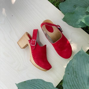 May include: A pair of red suede clogs with wooden heels and a buckle closure. The clogs are on a light wood surface with green leaves in the background.