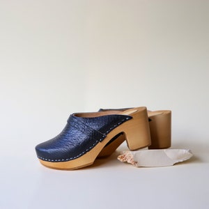 May include: A pair of black leather clogs with a textured finish, featuring wooden soles and heels. The clogs have a rounded toe and are accented with white stitching. A small, off-white object rests near the shoes.