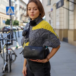 Può includere: Una donna indossa un maglione a righe grigio e giallo con una borsa a tracolla nera. La borsa ha un motivo a pelle di serpente e il nome del marchio "KULIK" è visibile sul davanti.
