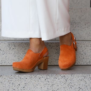 May include: A pair of orange suede clogs with wooden heels and a strap around the ankle. The clogs are worn by a person wearing white pants.
