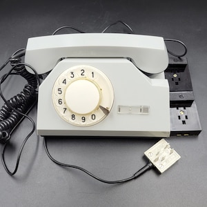 May include: A vintage rotary dial phone with a gray body and a black coiled cord. The phone has a black dial with white numbers 1 through 9 and a white dial with the numbers 0, 2, 3, 4, 5, 6, 7, 8, and 9. The phone has a black receiver and a black handset.