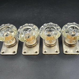 May include: Four vintage clear glass door knobs with a floral design. Each knob is mounted on a rectangular metal backplate with screw holes. The knobs have a brass-coloured base and are arranged in a row.