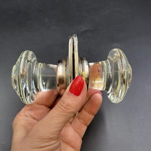 May include: Clear glass door knob with a ribbed design. The knob has a metal spindle and mounting hardware. The glass is transparent, and the metal has a tarnished appearance. The knob is held in a hand with red nail polish.