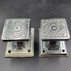May include: Two silver door knobs with a decorative design. The knobs are square shaped and have a white base.