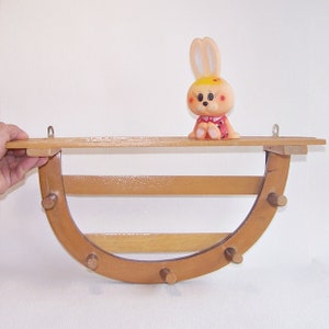 Puede incluir: Una estantería de madera con fondo curvo y ganchos. La estantería tiene un acabado marrón claro y está montada en una pared. Un pequeño conejito de juguete de plástico está sentado en la estantería.