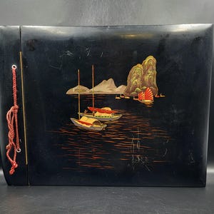May include: A black lacquered photo album featuring a painted scene of a seascape with boats and mountains. The album has a red cord closure.