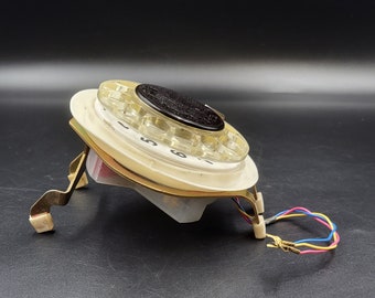 May include: A vintage rotary phone dial with a white plastic base and gold-colored metal accents. The dial has black numbers and a black center button. The phone is on a black surface.