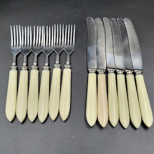 May include: A set of vintage silverware, including forks and knives. The forks have four tines and cream-colored handles. The knives have a silver blade and cream-colored handles. The silverware is arranged on a dark surface.