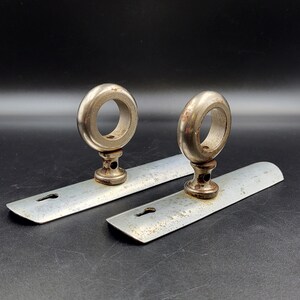 May include: Two silver-toned metal drawer pulls featuring a flat, rectangular backplate with a keyhole cutout and a circular ring handle attached to a central pedestal. The metal surfaces show signs of oxidation and wear, with visible rust spots.