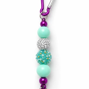 May include: A keychain featuring a purple metal carabiner attached to a series of beads. The sequence includes two small metallic purple beads, two smooth mint green round beads, one white bead with black scribbled patterns, and a textured teal bead.