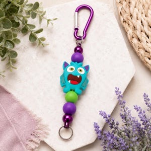 May include: A keychain featuring a teal monster character bead with a wide mouth and white teeth, accented by purple and lime green spherical beads, a metallic purple carabiner clip at the top, and a silver metal ring at the bottom.