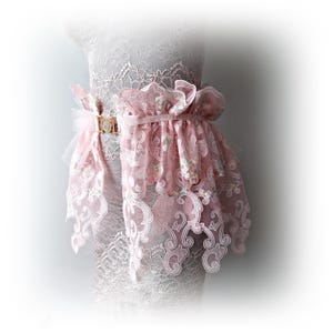 May include: A pink lace belt with floral embroidery and a gold buckle. The belt features a layered design with delicate, flowing fabric and intricate scrollwork patterns. The belt is displayed on a mannequin.