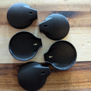 Puede incluir: Cinco tapones de botella negros en forma de lágrima con un pequeño agujero en la parte superior, dispuestos sobre una superficie de madera. Los tapones tienen un acabado liso y mate y un borde ligeramente elevado.