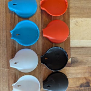 以下が含まれることがあります： 青、オレンジ、白、黒の、水滴型のカラフルなボトルストッパーのコレクション。各ストッパーには小さな穴と湾曲したハンドルが付いています。黒いストッパーには「JCI」の文字が印刷されています。