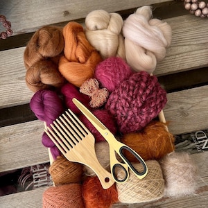 Peut inclure: Une collection de laine peignée et de fil dans des tons marron, orange, rose et bordeaux, accompagnée d'un peigne à laine en bois et de ciseaux dorés à poignées noires, le tout disposé dans un plateau rectangulaire en bois.
