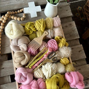 Puede incluir: Una bandeja de madera llena de hilo de colores para tejer. El hilo es en tonos de rosa, amarillo y beige. Hay un peine de madera, tijeras y algunas otras herramientas en la bandeja.