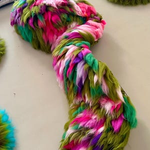 Könnte beinhalten: Ein langes, strukturiertes Faser-Kunstwerk mit einer lebendigen Mischung aus langen, zotteligen Strähnen in Olivgrün, Pink, Lila und Hellrosa. Die Fasern sind dicht gepackt und geschichtet, was ein voluminöses, mehrfarbiges Erscheinungsbild erzeugt.