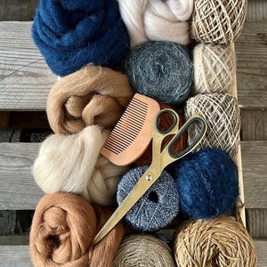 Peut inclure: Un plateau en bois rempli de fournitures artisanales. Il contient des pelotes de laine bleues, marron et beiges, ainsi que de la laine en mèches. Un peigne en bois et des ciseaux dorés suggèrent un intérêt pour les arts textiles.