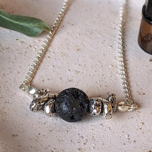以下が含まれることがあります： 黒い溶岩のビーズと銀のチャームが付いた銀のチェーンネックレス。黒いキャップが付いた小さな茶色のガラス瓶が白いセラミックの皿の上に置かれています。