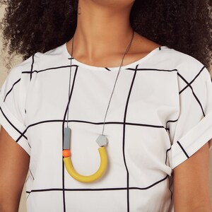 May include: A woman with curly black hair is wearing a white and black checked shirt with a yellow, orange and grey necklace.