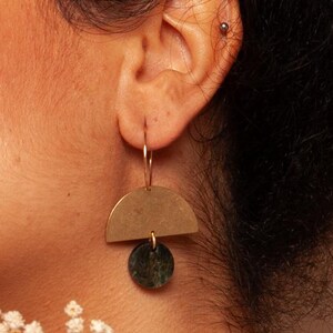 May include: A close-up of a person's ear wearing a pair of brass earrings. Each earring features a half-circle shape above a dark green circular stone. The earrings hang from a gold hoop. The background is a soft, warm color.