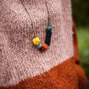 May include: A colourful wooden bead necklace with yellow, orange, blue, green and black beads. The necklace is worn by a person wearing a pink and orange jumper.
