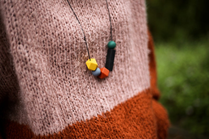 May include: A colourful wooden bead necklace with yellow, orange, blue, green and black beads. The necklace is worn by a person wearing a pink and orange jumper.