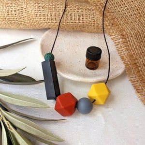 May include: A black cord necklace with colourful silicone beads. The necklace features a black rectangular bead, a red geometric bead, a grey round bead, and a yellow geometric bead. A small brown glass bottle with a black lid is also pictured.