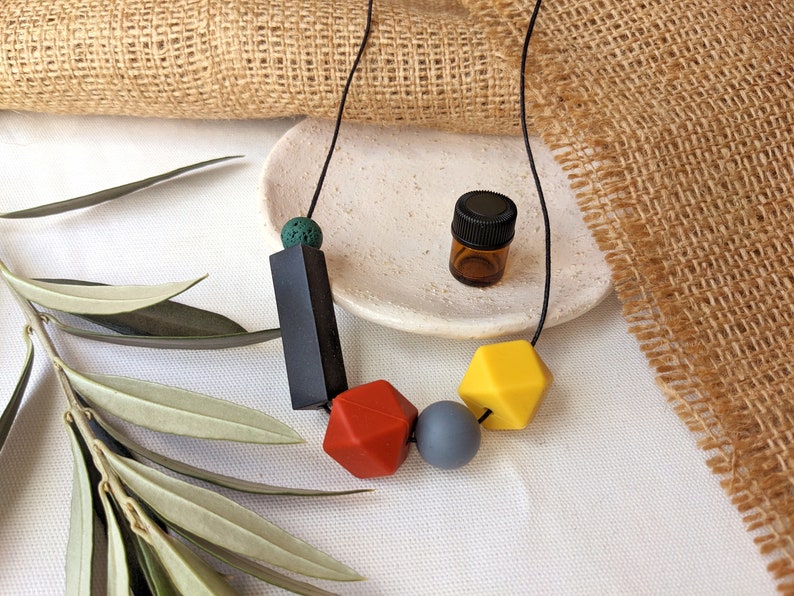 May include: A black cord necklace with colourful silicone beads. The necklace features a black rectangular bead, a red geometric bead, a grey round bead, and a yellow geometric bead. A small brown glass bottle with a black lid is also pictured.