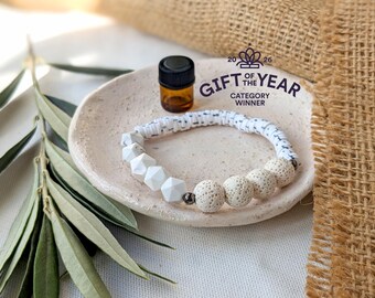 May include: A white and gray beaded bracelet with white geometric beads and porous beige beads. A small amber glass bottle with a black cap sits next to the bracelet on a small, light-colored dish. The text "GIFT OF THE YEAR CATEGORY WINNER" is visible.