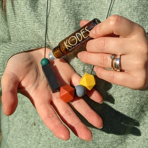 May include: A brown glass bottle with a black label that says "KODES" and "ORGANIC ESSENTIAL OIL FOR RELAXATION". The bottle is on a black cord with green, black, red, grey, and yellow silicone beads.