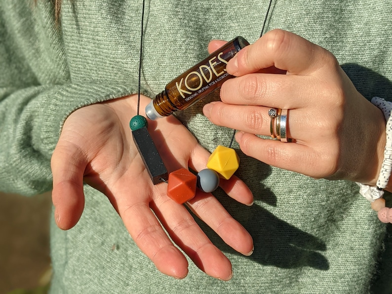 May include: A brown glass bottle with a black label that says "KODES" and "ORGANIC ESSENTIAL OIL FOR RELAXATION". The bottle is on a black cord with green, black, red, grey, and yellow silicone beads.