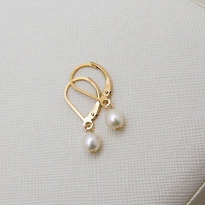 May include: Gold-coloured leverback earrings, each with a pear-shaped pearl. The earrings are presented on a white, textured surface. The leverback design ensures secure wear. A classic jewellery item.
