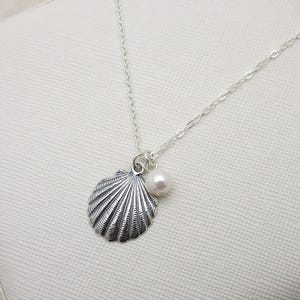 May include: A silver seashell pendant necklace with a small pearl charm. The pendant has a detailed, ridged design. The necklace is displayed on a white, textured surface, suggesting a jewelry presentation.