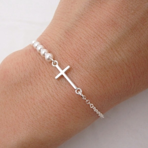 May include: A silver bracelet featuring a small cross pendant and a chain. The bracelet also includes a series of small, round beads. The bracelet is shown on a wrist.