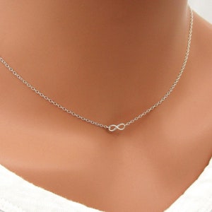 May include: A delicate silver necklace with a small infinity symbol pendant. The necklace is on a model's neck and is a simple, minimalist design.