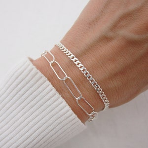 May include: Two silver bracelets on a wrist. One bracelet is a curb chain and the other is a chain with oval links.