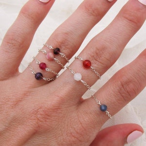 May include: A hand model wearing five delicate silver rings with small gemstone beads. The rings feature a variety of colors, including red, pink, white, and blue.