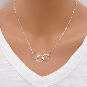 May include: A silver necklace with three interlocking circles pendant. The necklace is on a mannequin wearing a white shirt.
