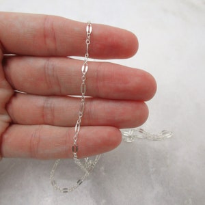 May include: A silver chain necklace with a delicate, flat oval link design. The chain is shown draped over a hand.