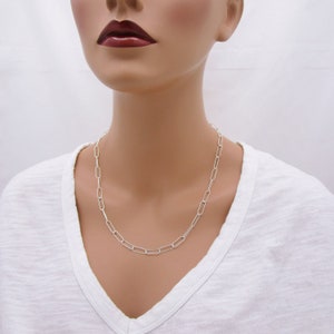 May include: A silver-colored chain necklace with elongated rectangular links. The necklace is worn around the neck and rests on a white v-neck shirt. The links are evenly spaced and create a simple, elegant design.